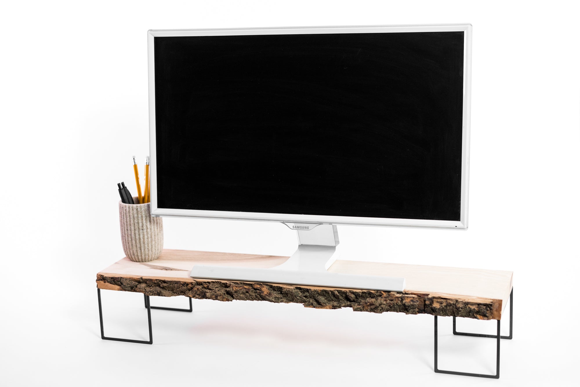 Computer monitor on a  wooden stand with a natural bark edge, supported by minimalist metal legs. A ceramic holder with pencils is placed on the left side of the stand.